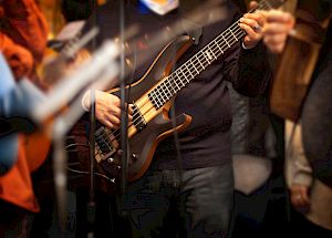 A person playing a bass guitar during a live performance, surrounded by other musicians and equipment in a dimly lit setting.